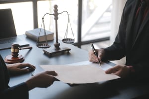 Zwei Personen bei einer Vertragsunterzeichnung im Anwaltsbüro, mit Justitia-Waage und Richterhammer im Hintergrund.