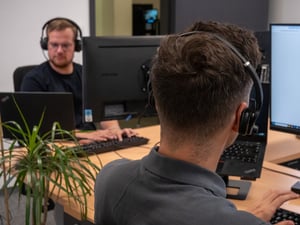 Zwei Mitarbeiter mit Headsets arbeiten an ihren Schreibtischen mit mehreren Monitoren in einem Büro.