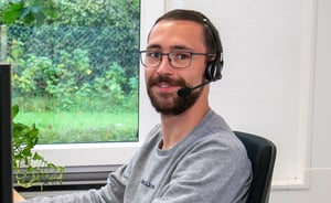 Mitarbeiter mit Headset sitzt an einem Schreibtisch und schaut lächelnd in die Kamera