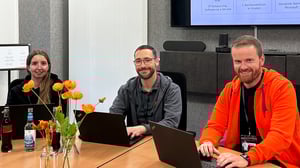 Drei lächelnde Personen sitzen gemeinsam an einem Konferenztisch mit Laptops und Getränken