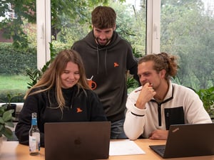 Drei Personen sitzen und stehen an einem Tisch mit Laptops und Unterlagen