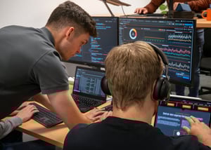 Zwei Personen sitzen an Schreibtischen mit mehreren Monitoren und Laptops. Auf den Bildschirmen sind Diagramme, Tabellen und Benutzeroberflächen zu sehen